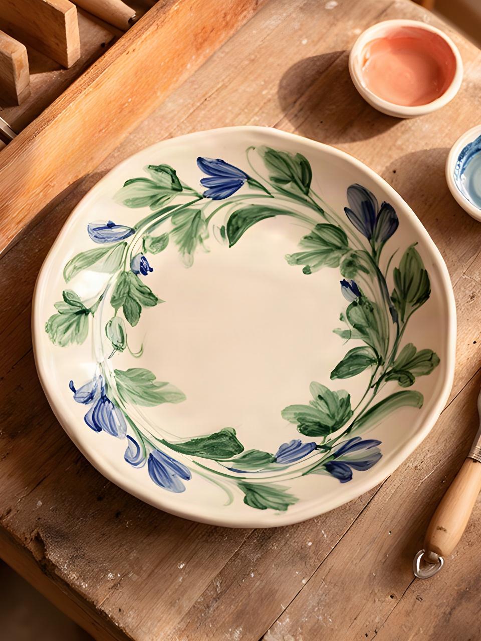 Piatto in maiolica dipinto a mano con foglie verdi e dettagli blu, appoggiato su un banco di legno
