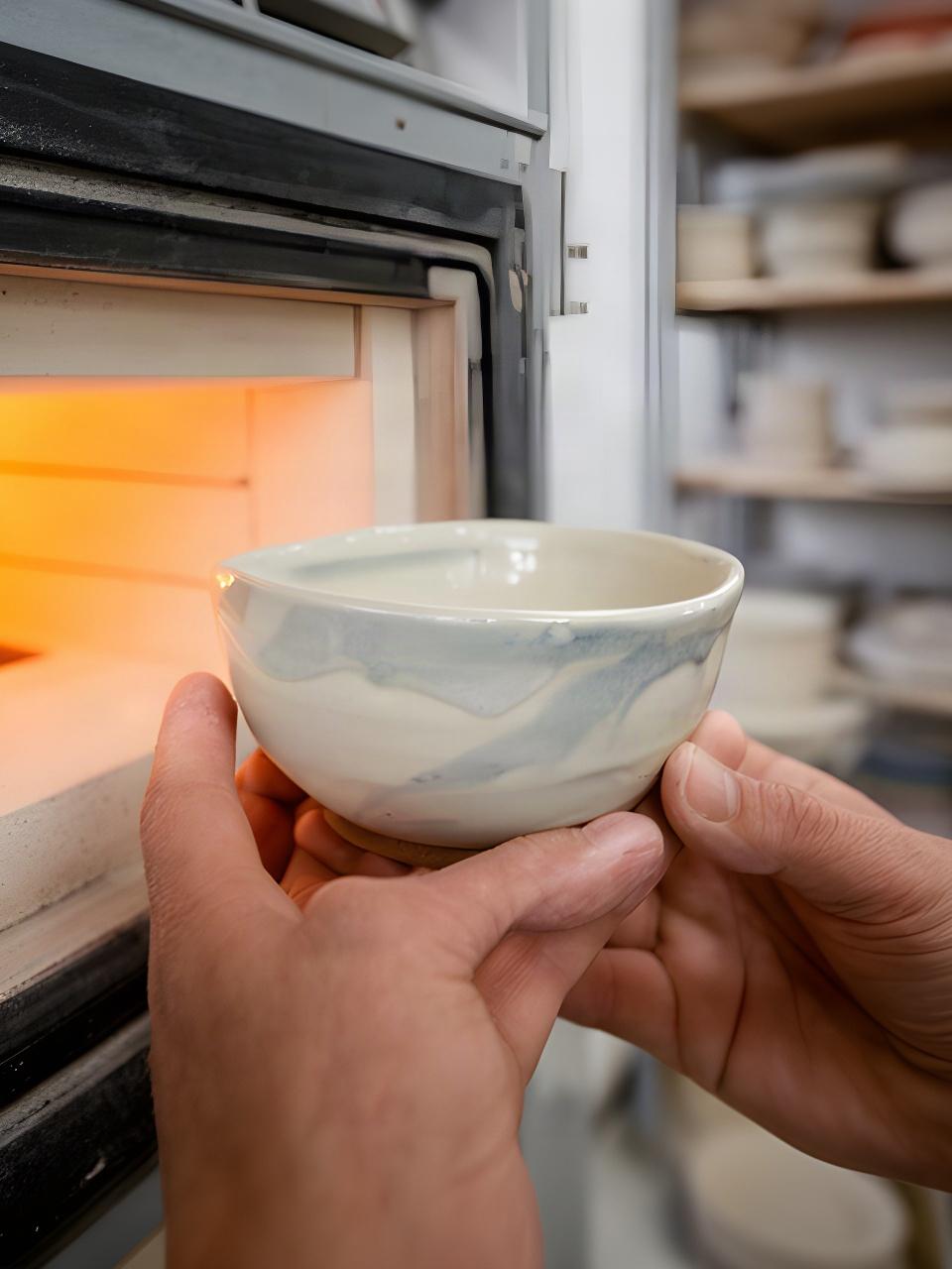 Mani che sostengono una ciotola smaltata accanto a un forno ceramico nello studio artigianale